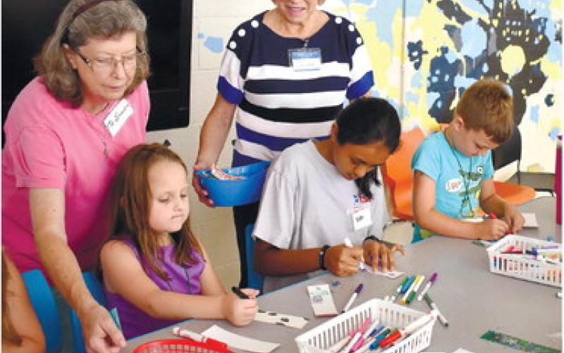 Volunteers Sandra Gunn (left), and Laura Hotzler watch as campers color bookmarks for veterans.  LENA HENSLEY/Staff