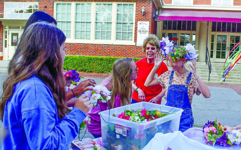Abigail Hildreth tries on a flower crown she just made, Jade Mullis is having a close look to make sure it fits, and volunteer Janet Kelhoffer (behind them) is talking to the other girls working on their flower crowns.