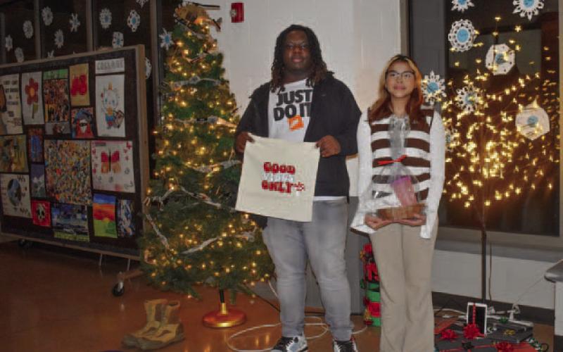 Posing in front of Christmas trees representing PCHS student organizations, graphic design students Scarlett Monje (11th grade) and Tavion Davis (12th) showcase items they designed in class and sold at the silent auction.