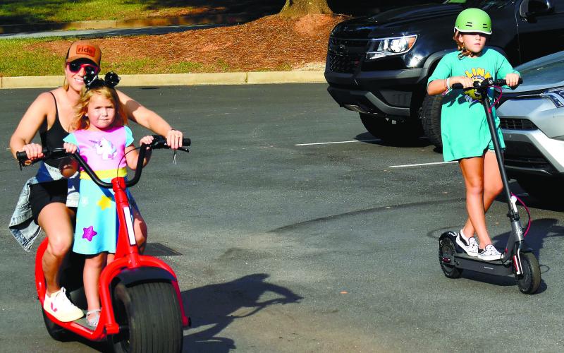 Angela Sauter gives younger daughter Elowen a ride while older sister Hadley rides alongside on an electric scooter.