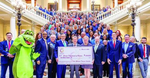 Gov. Brian Kemp (front center) accepts a symbolic $5 billion check from Amanda Dyson-Thornton, executive director of the Georgia Association of Convention &amp; Visitors Bureaus. MMCVB Executive Director Jennifer Rosa León is at top right of the main staircase before it turns right. CONTRIBUTED