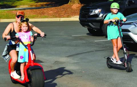 Angela Sauter gives younger daughter Elowen a ride while older sister Hadley rides alongside on an electric scooter.